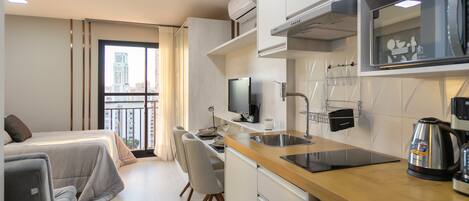 A compact hotel room with a kitchenette, a bed, a dining area, and a large window offering a city view.