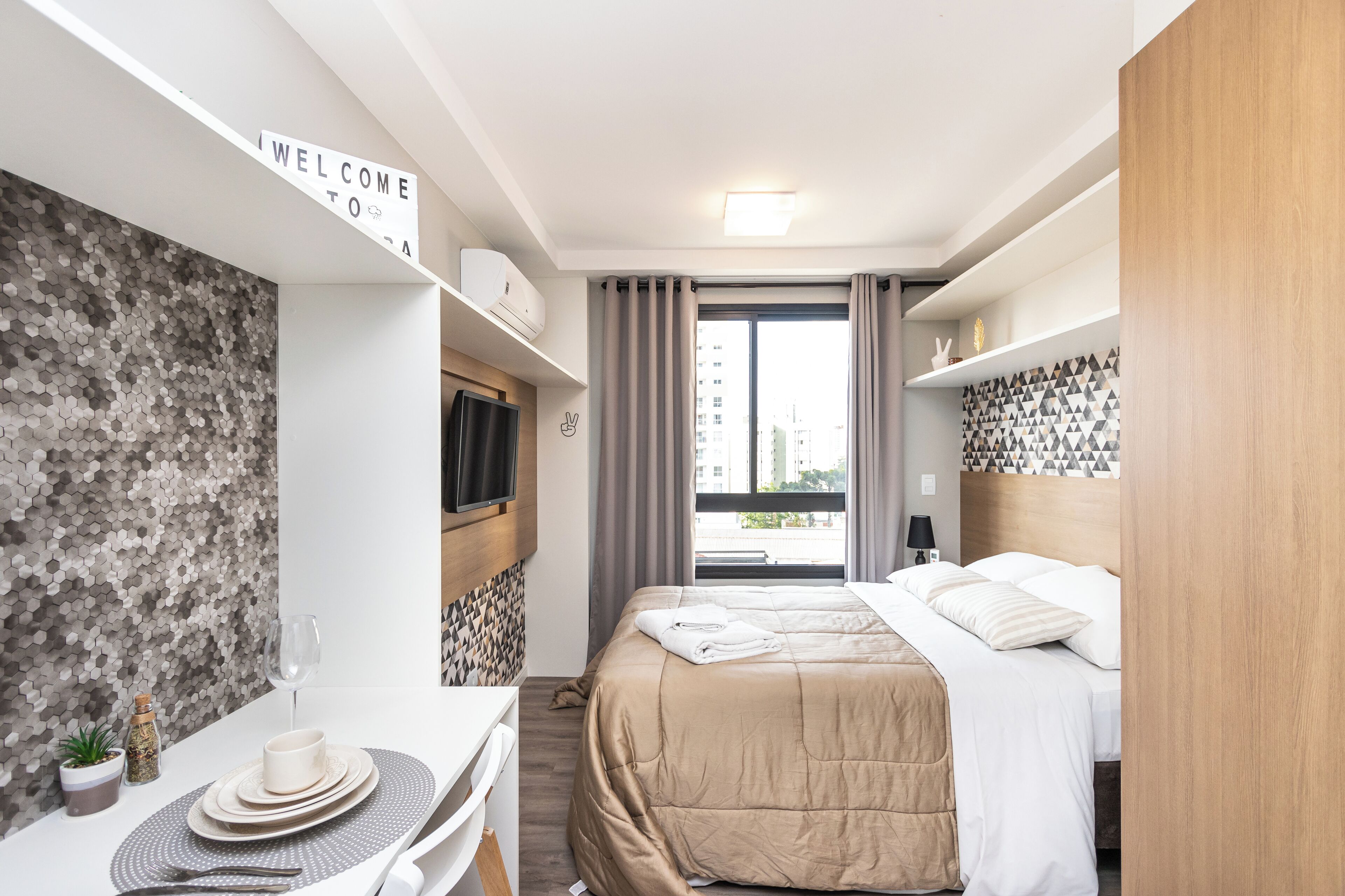 A modern hotel room with a bed, a TV, a dining table with a placemat, a "WELCOME TO" sign, and a city view through the window.