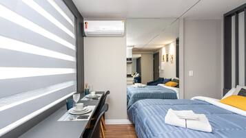 Ett kompakt hotellrum med en enkel säng, ett litet skrivbord med en stol och ett fönster med persienner.