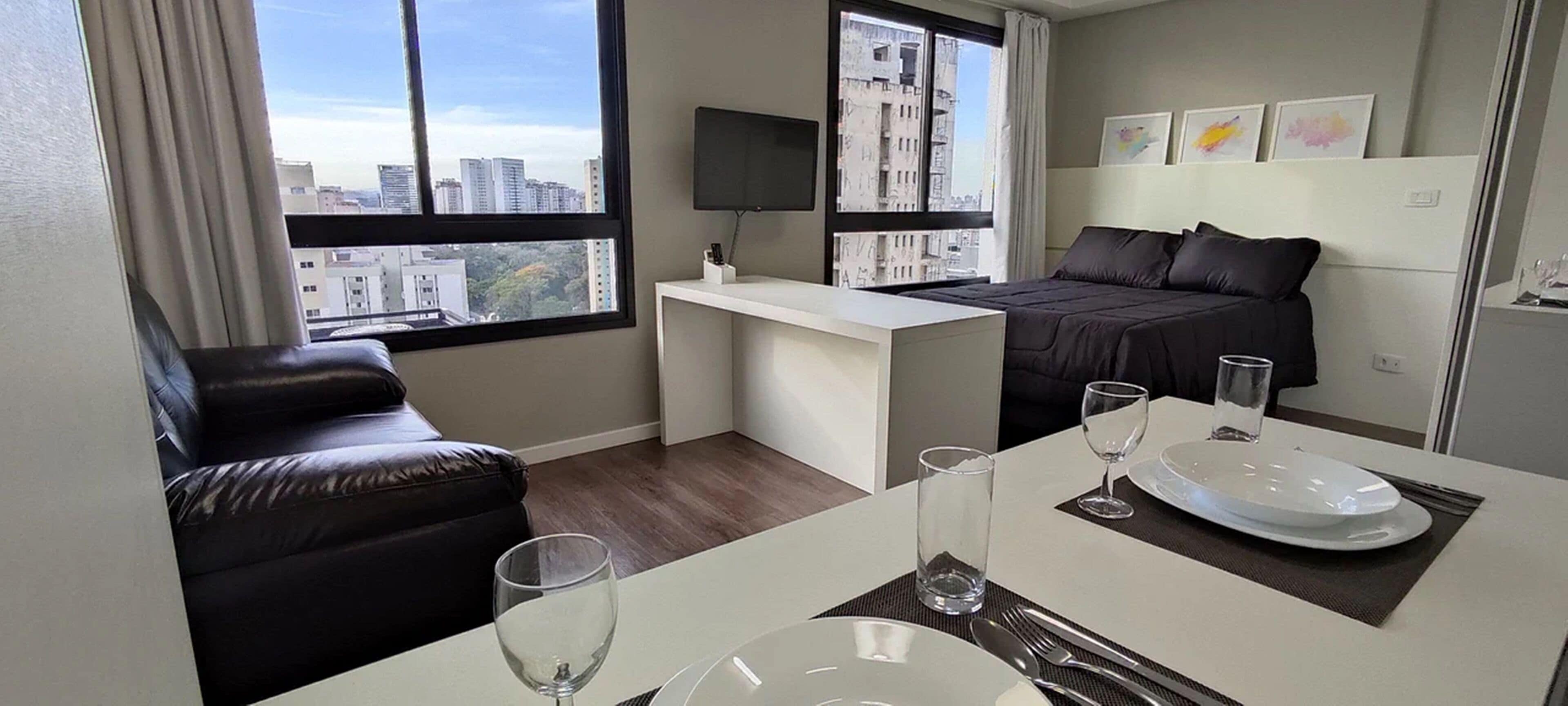 Ein modernes Hotelzimmer mit einem Bett, einem Schreibtisch, einem Ledersofa, einer Essecke mit Tisch für zwei Personen und großen Fenstern mit Blick auf die Stadt.