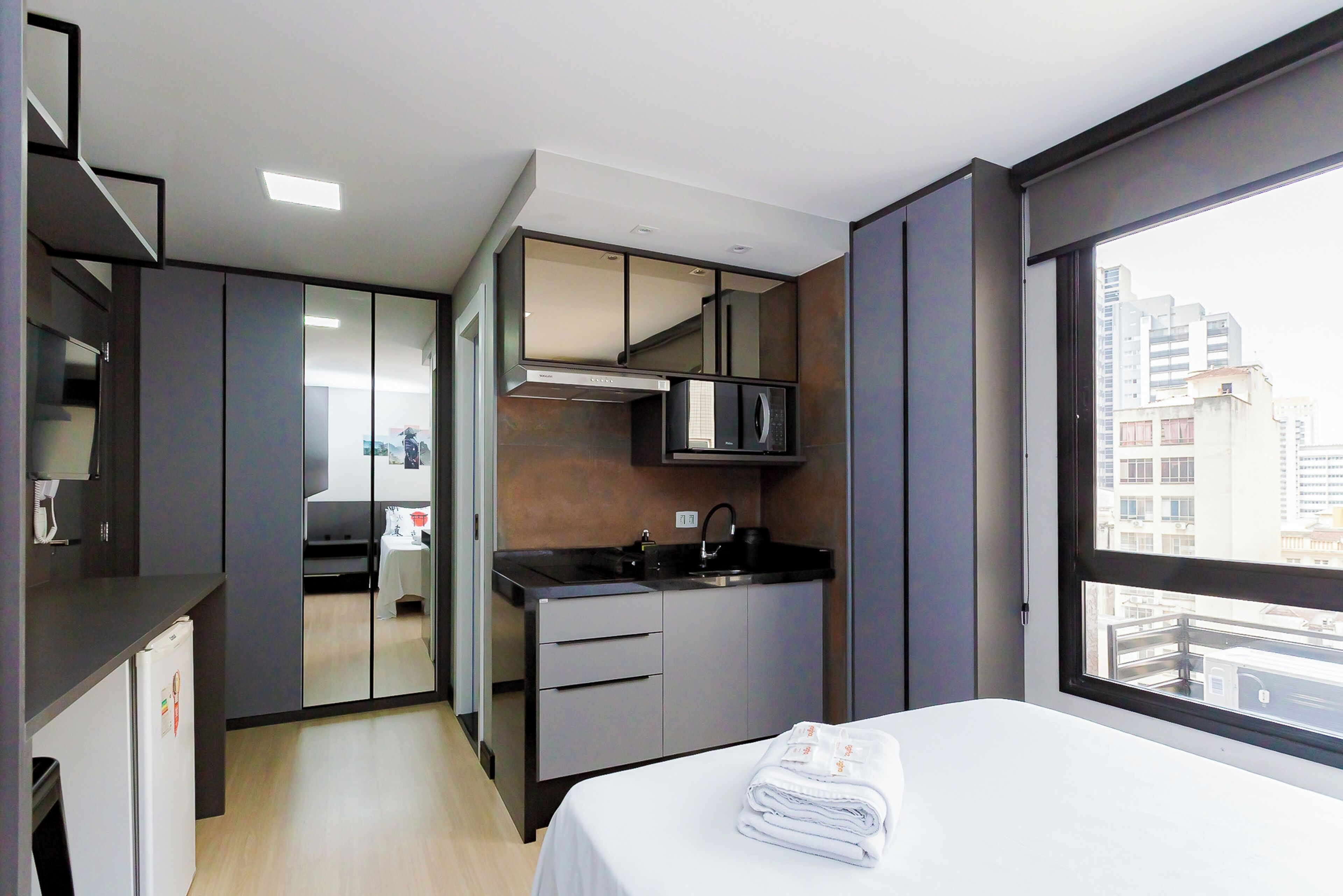 Une chambre d’hôtel moderne équipée d’un lit, d’une petite kitchenette et d’une grande fenêtre donnant sur la ville.