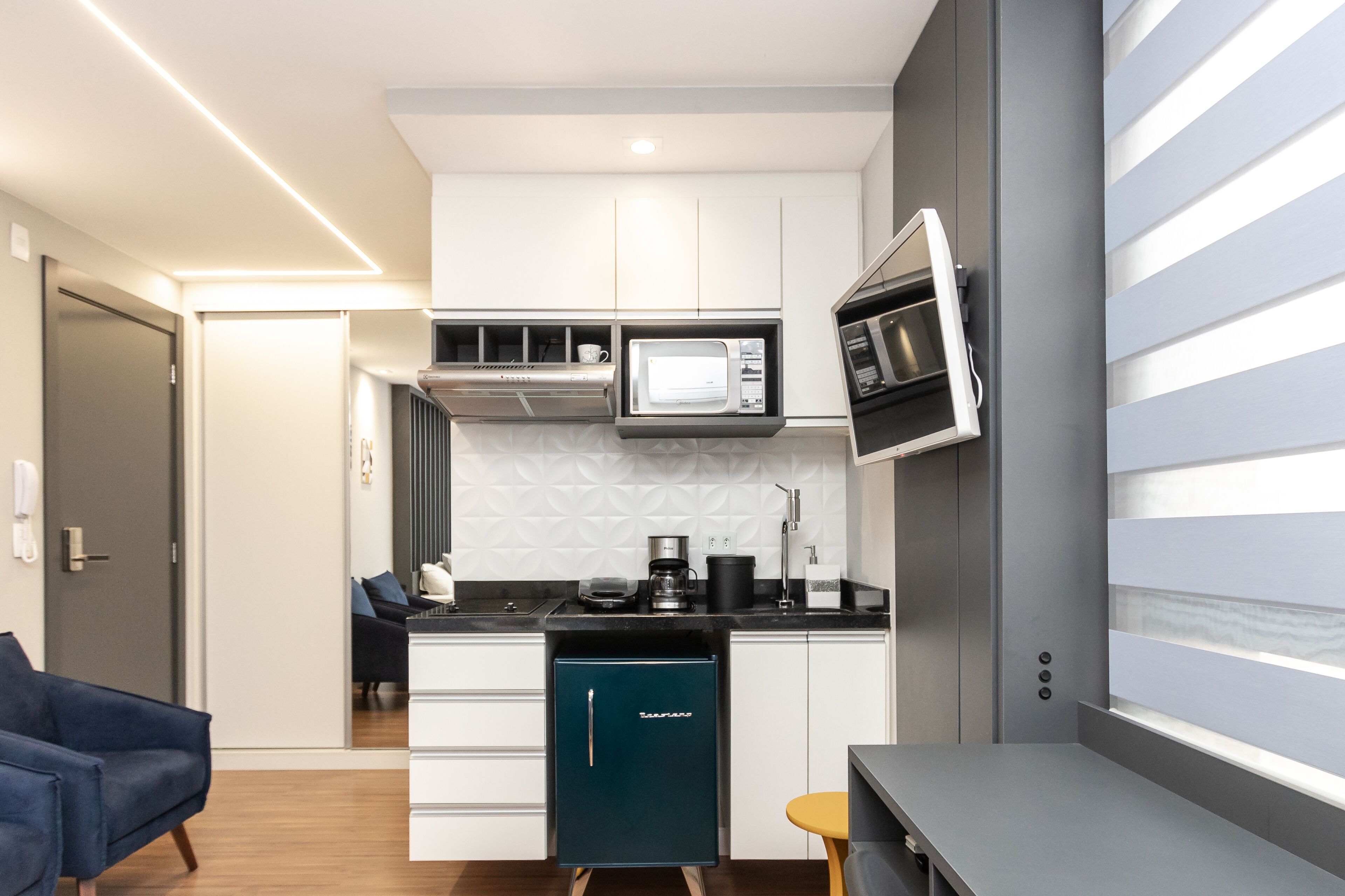 Cocina compacta con armarios blancos, microondas, cafetera y refrigerador pequeño.