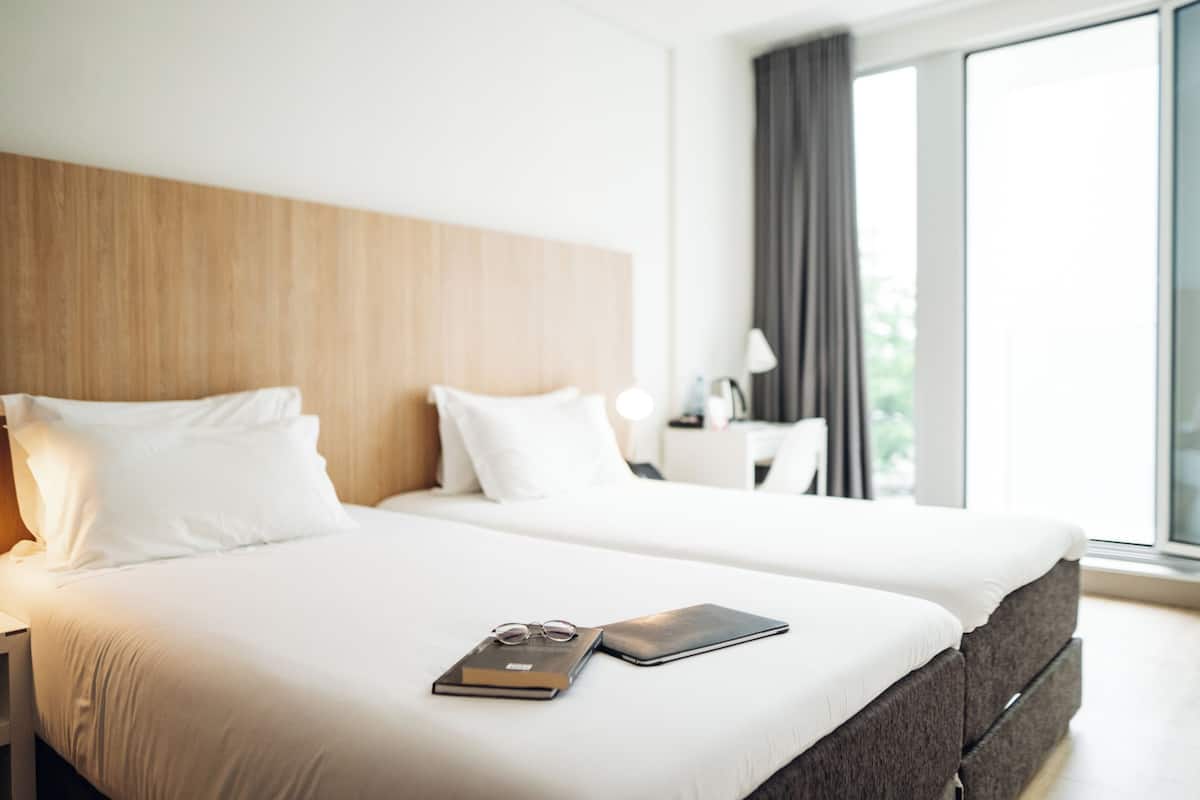 Hotelkamer met twee bedden, een houten hoofdbord, wit beddengoed, een nachtkastje met een lamp, en uitzicht op groen door grote ramen.