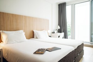 Hotelkamer met twee bedden, een houten hoofdbord, wit beddengoed, een nachtkastje met een lamp, en uitzicht op groen door grote ramen.