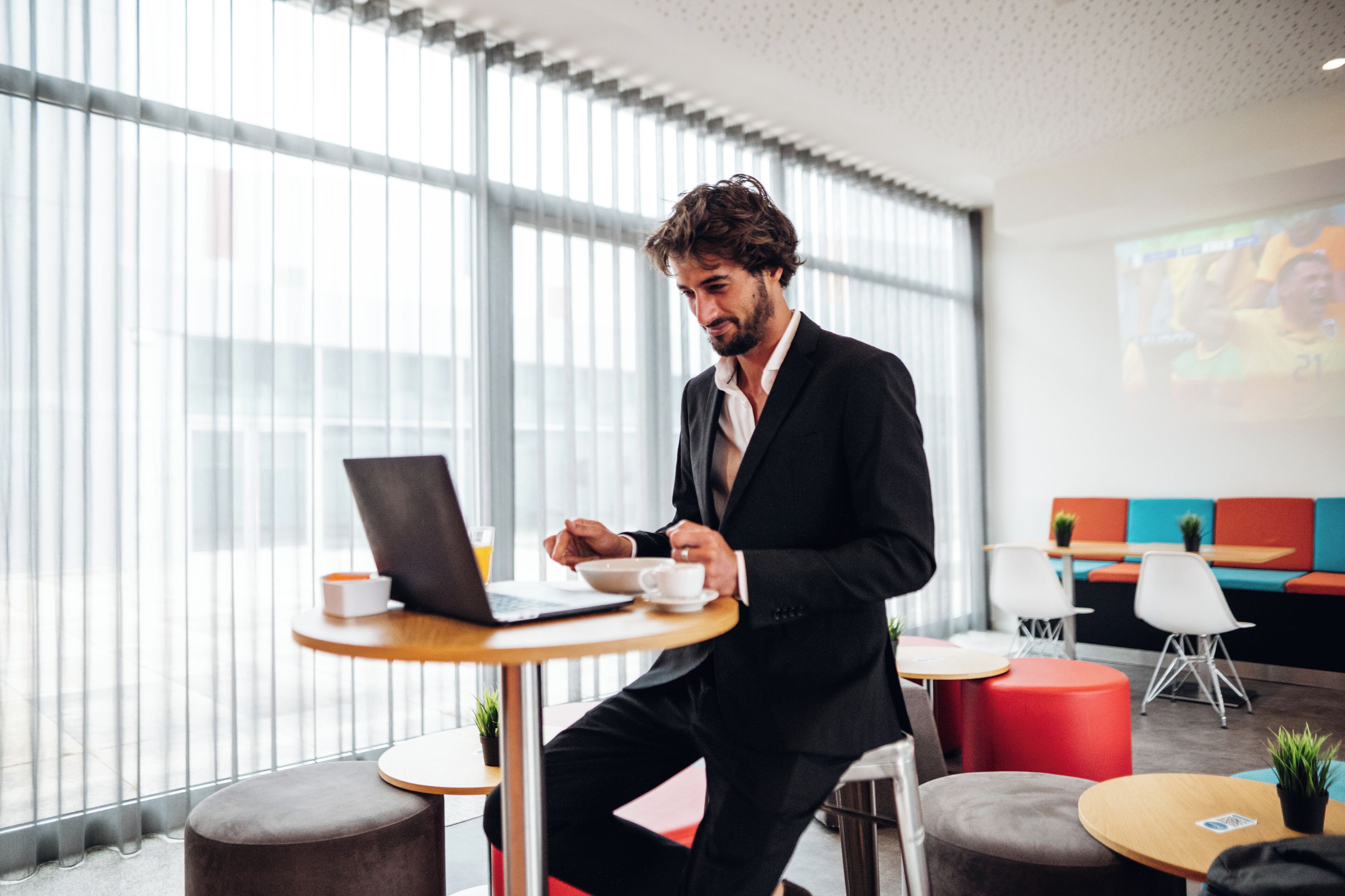 Ein Mann in einem schwarzen Anzug arbeitet an einem Laptop an einem Holztisch in einem modernen Büro mit großen Fenstern und bunten Sitzbereichen.