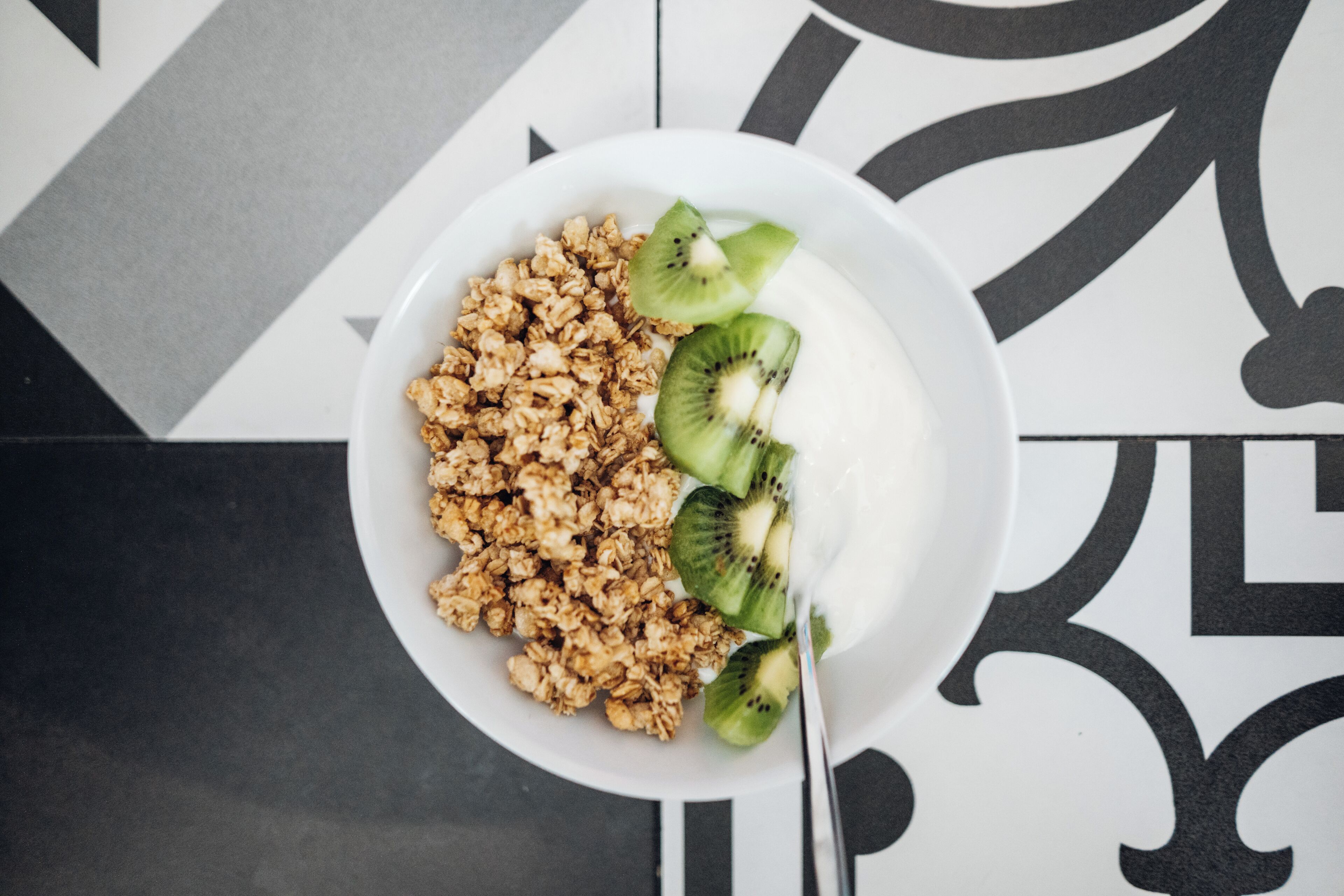 A white plate with granola, kiwi slices, and yogurt on a black and white patterned floor.