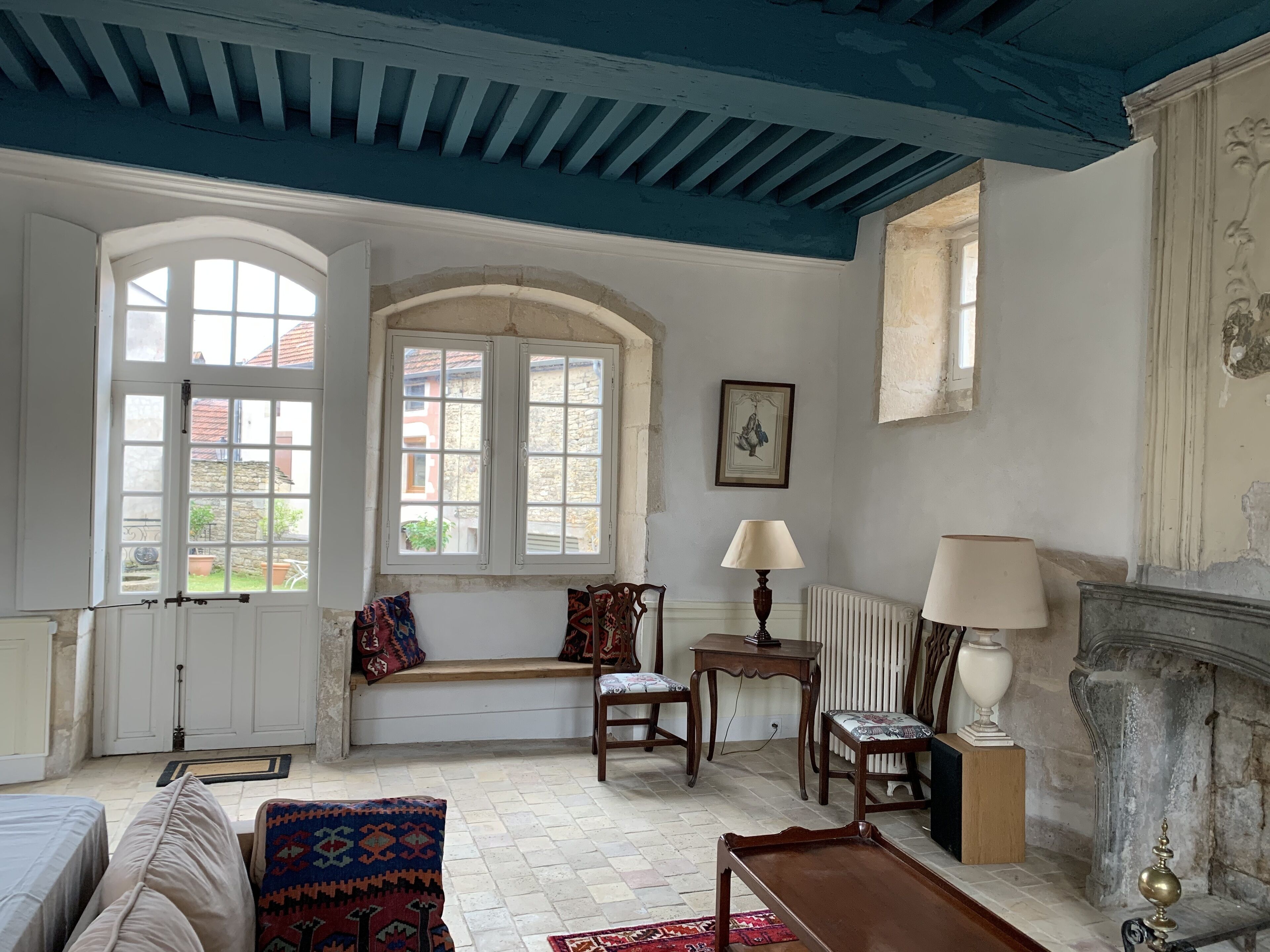 A room with a high ceiling, white walls, and a blue painted ceiling. There are two windows with white frames, a fireplace, and a table with a lamp.