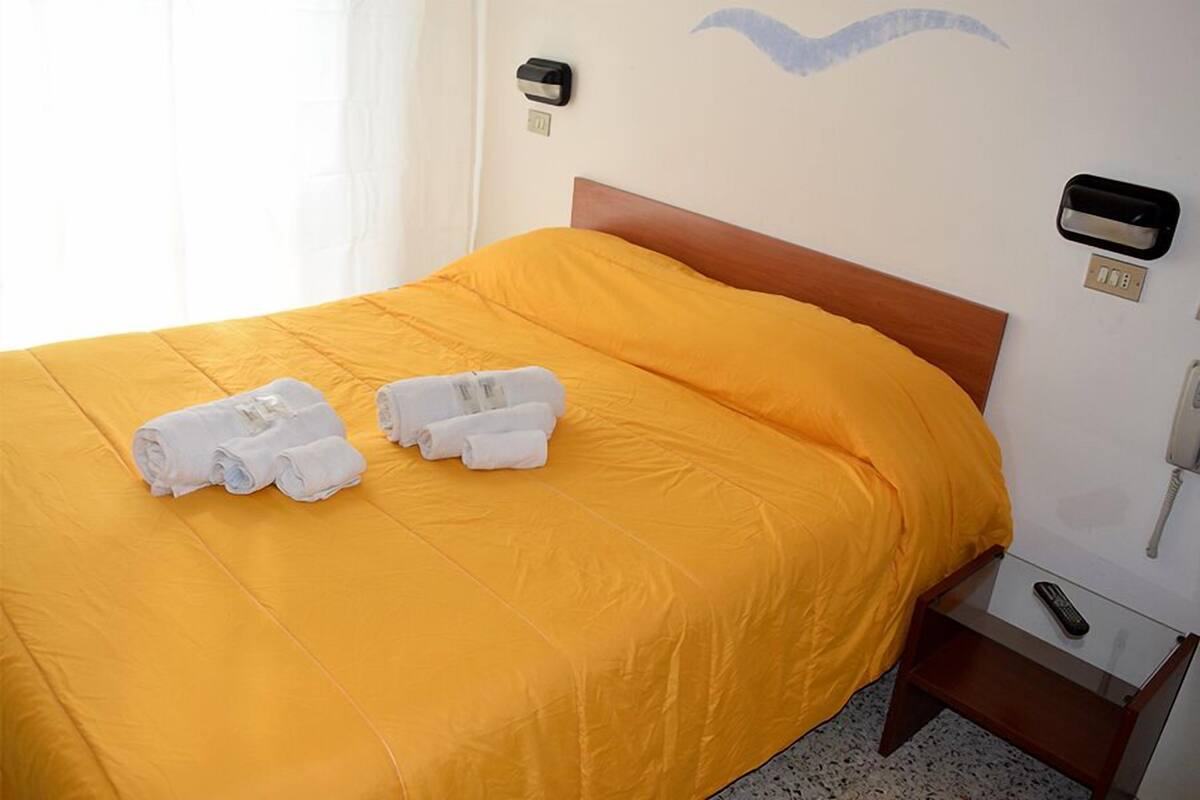 A hotel room with a bed covered in yellow bedding, two rolled white towels on the bed, a wooden headboard, a bedside table with a phone, and a wall-mounted light switch.