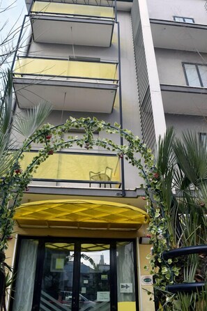 A building with a yellow awning and a glass door entrance, surrounded by greenery. - Hotel Criss (Rimini)