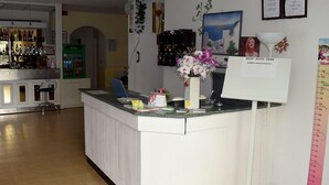Un mostrador de recepción de hotel con una computadora, un panel de menú y una zona de bar al fondo.