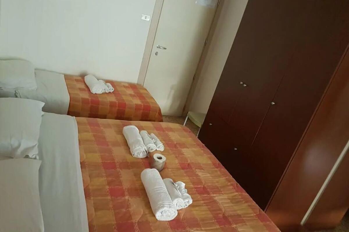 A hotel room with two beds, each with a checkered bedspread and rolled towels. There is a wardrobe and a wall-mounted telephone.