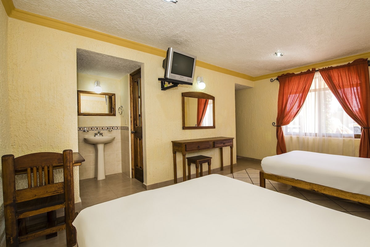 Ein Hotelzimmer mit zwei Betten, einem Holzstuhl, einem kleinen Tisch, einem Fernseher, einem Spiegel, einem Waschbecken und einem Fenster mit Vorhängen.