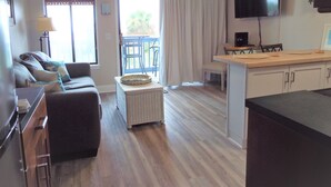 Un salon avec un canapé, une petite table, un téléviseur et un balcon équipé de chaises.