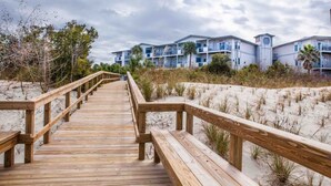 Une promenade en bois mène à un bâtiment situé en bord de mer, doté de nombreux balcons, avec une plage en arrière-plan.
