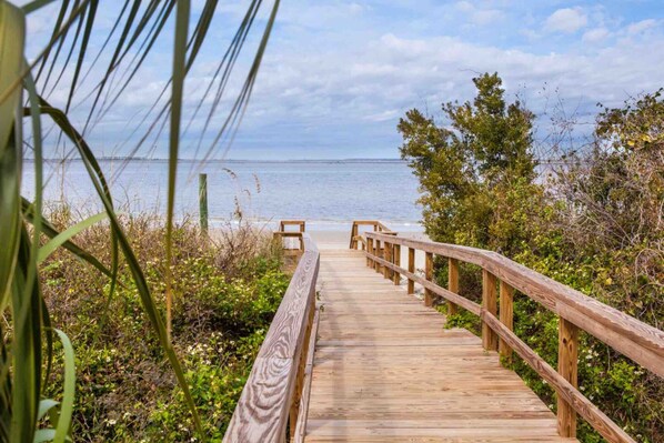 Walk along the boardwalk to the serene beach, surrounded by lush greenery and a clear sky.
