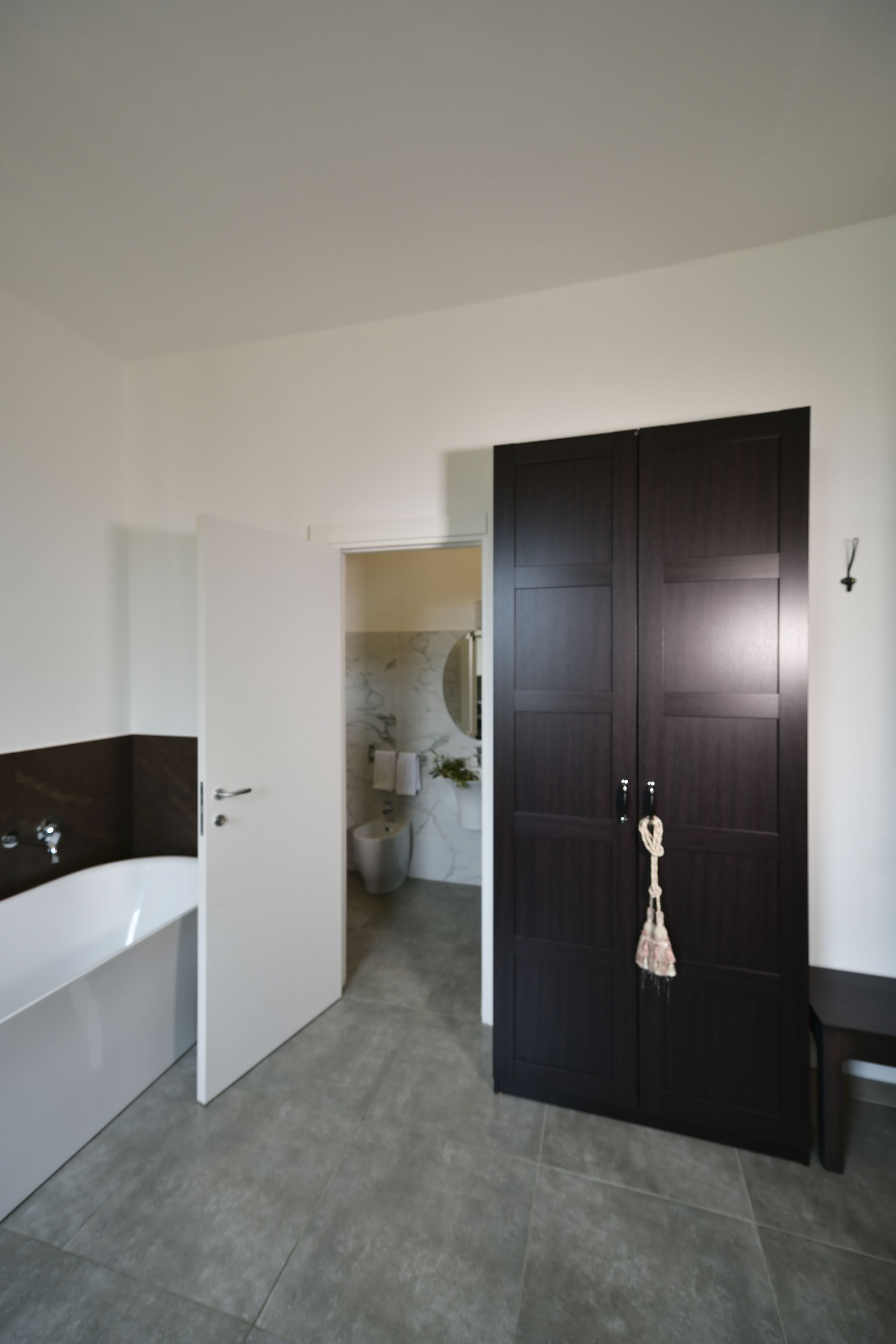 A modern bathroom with a dark wooden door, a white bathtub, and a marble wall.
