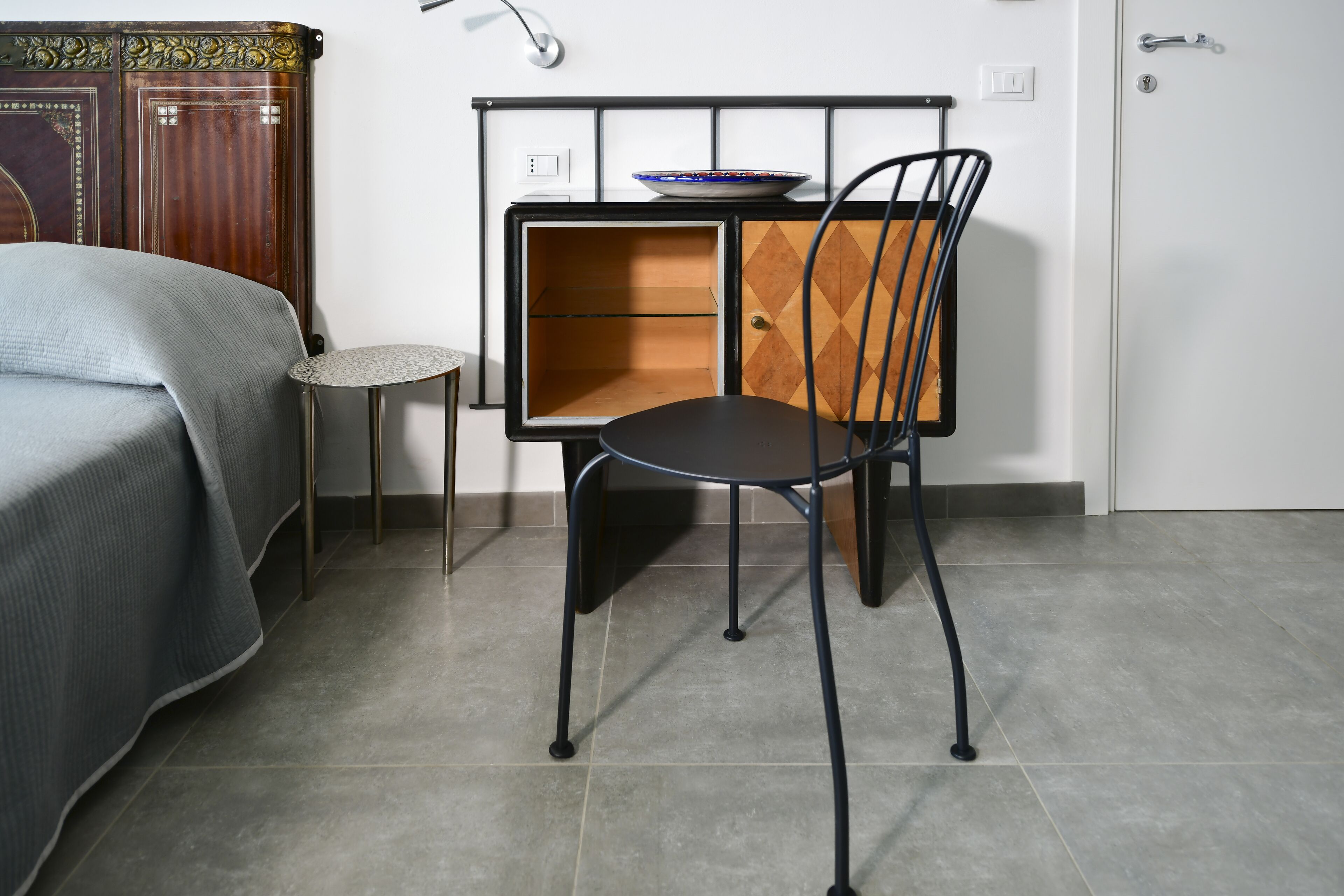 A room with a bed, a small round table, a chair, and a cabinet with a bowl on top.