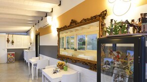 A dining area with white tables and chairs, a black display case with items, and a large mirror on the wall. - Inkantu B&B (Terrasini)