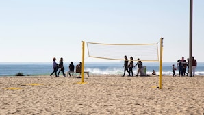 Ein Beachvolleyballfeld mit Netz, umgeben von Menschen und dem Meer im Hintergrund.