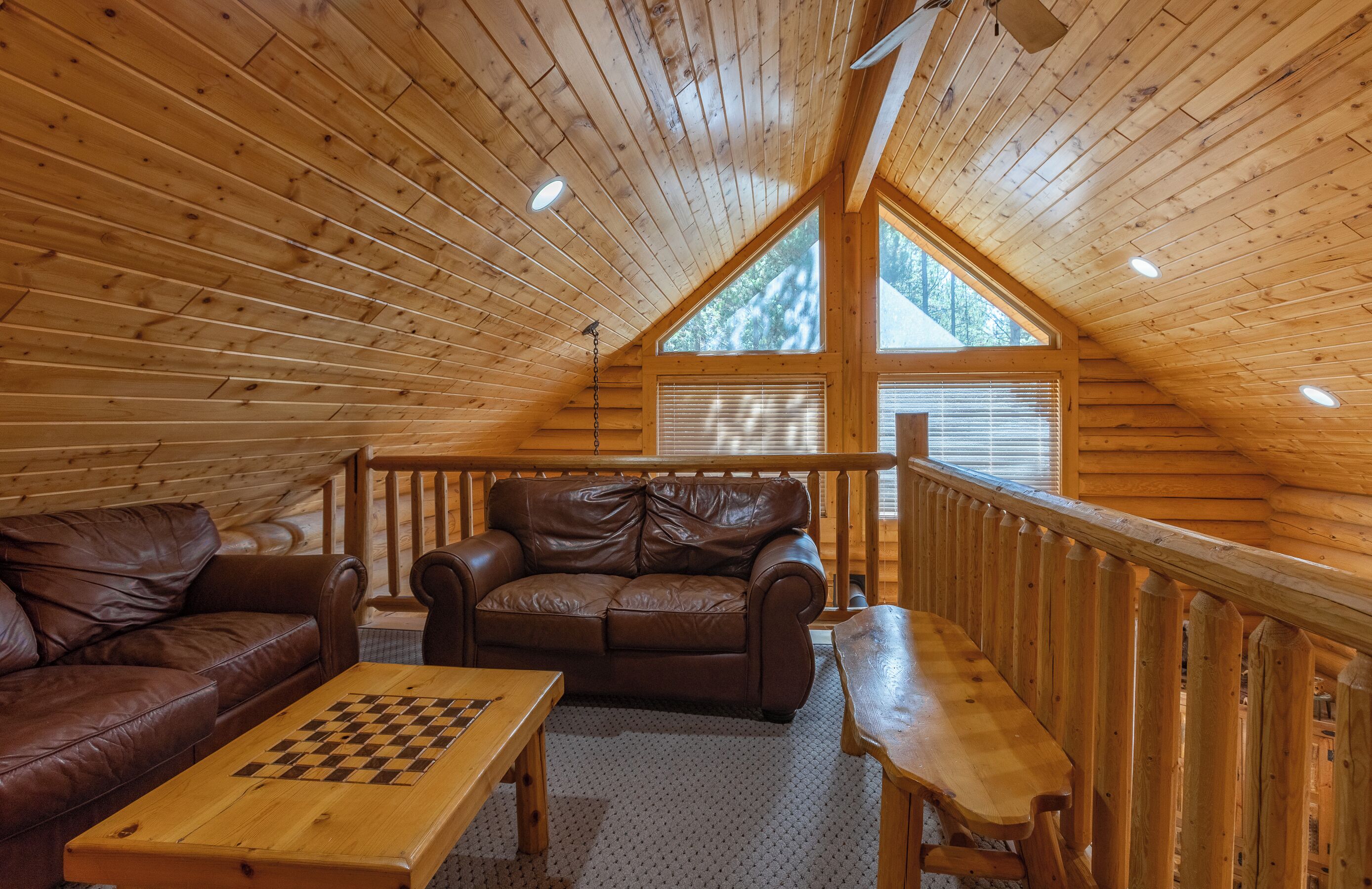 A cozy wooden cabin interior with a loft bedroom, a leather sofa, a wooden coffee table, and a dining table.