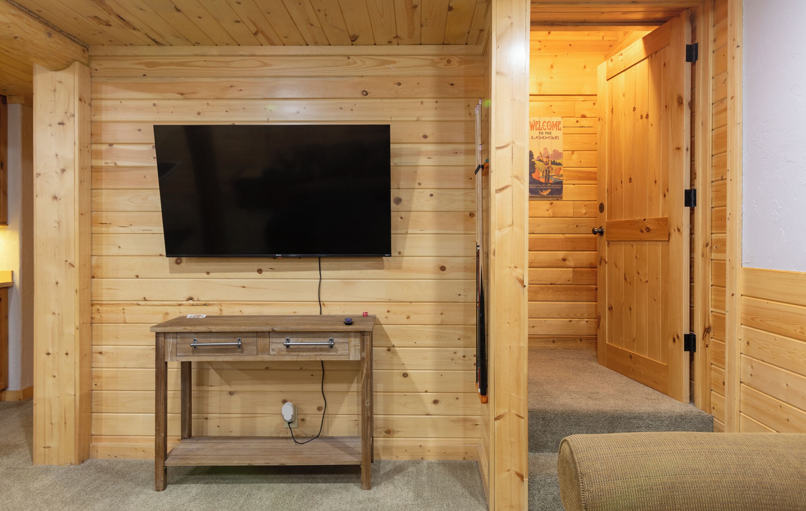 A wooden-paneled room with a flat-screen TV mounted on the wall, a wooden desk with drawers, and a door with a "Welcome" sign.