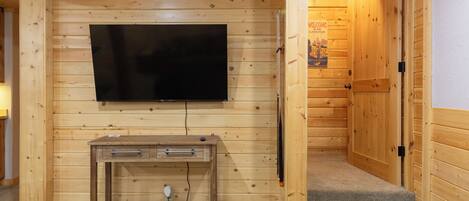 A wooden-paneled room with a flat-screen TV mounted on the wall, a wooden desk with drawers, and a door with a "Welcome" sign.