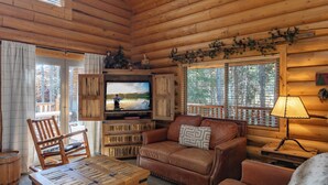 A cozy log cabin interior with a wooden ceiling, a television, leather furniture, and a window with a view of trees.