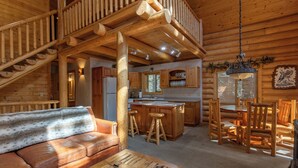 A log cabin interior with a wooden staircase, a kitchen with wooden cabinets, a dining area with wooden chairs, and a sofa with a fur throw.