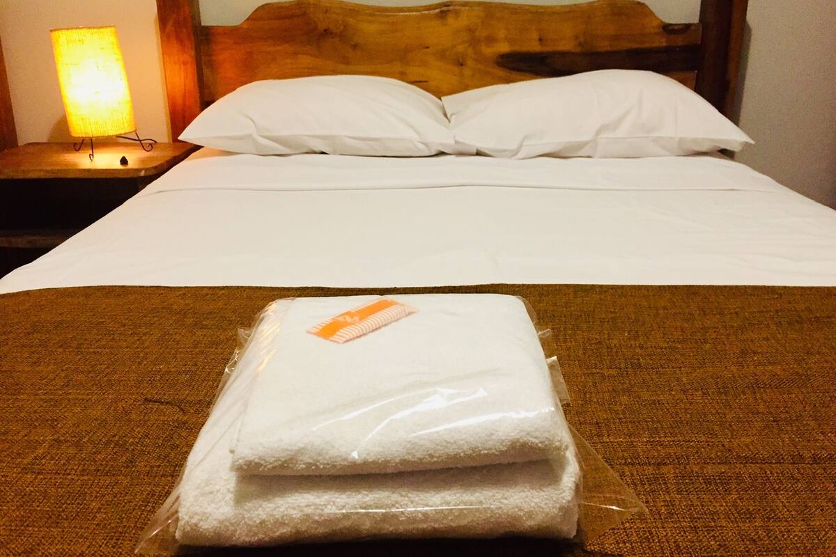 Una cama bien hecha con sábanas blancas, un cabecero de madera y una mesita de noche con una lámpara.