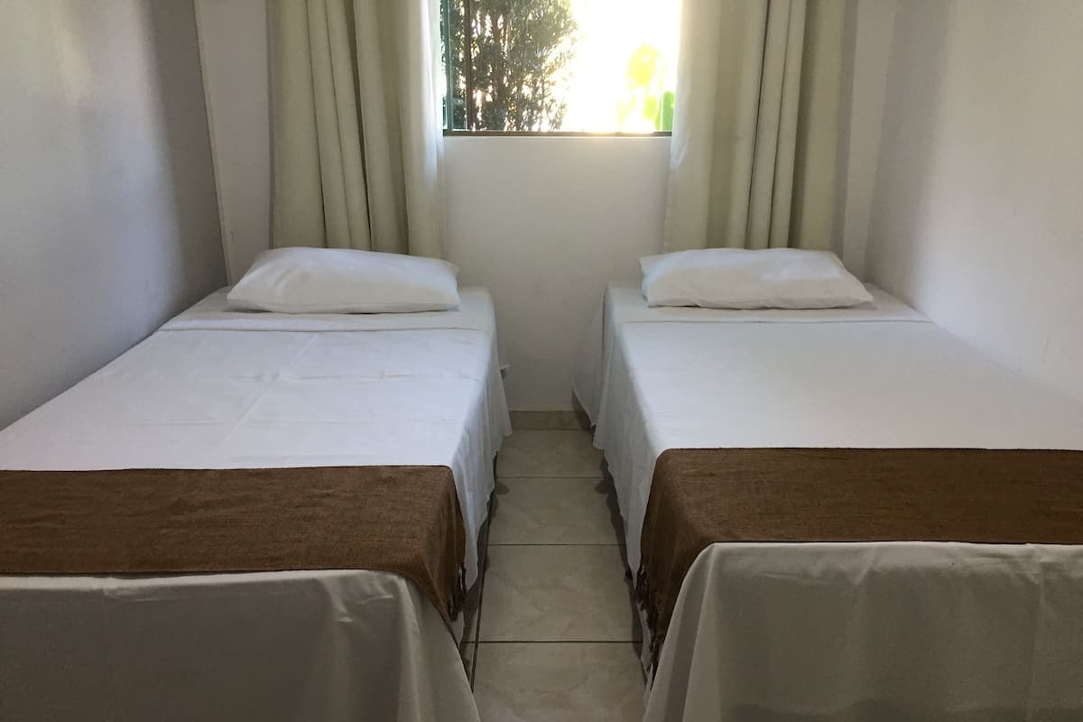 Dos camas individuales con sábanas blancas y cabeceros marrones, una ventana con cortinas y un suelo de baldosas.