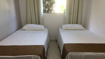 Duas camas de solteiro com roupa de cama branca e cabeceiras castanhas, uma janela com cortinas e piso de azulejos.