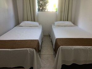 Deux lits simples avec du linge de lit blanc et des têtes de lit marron, une fenêtre avec des rideaux et un sol carrelé.