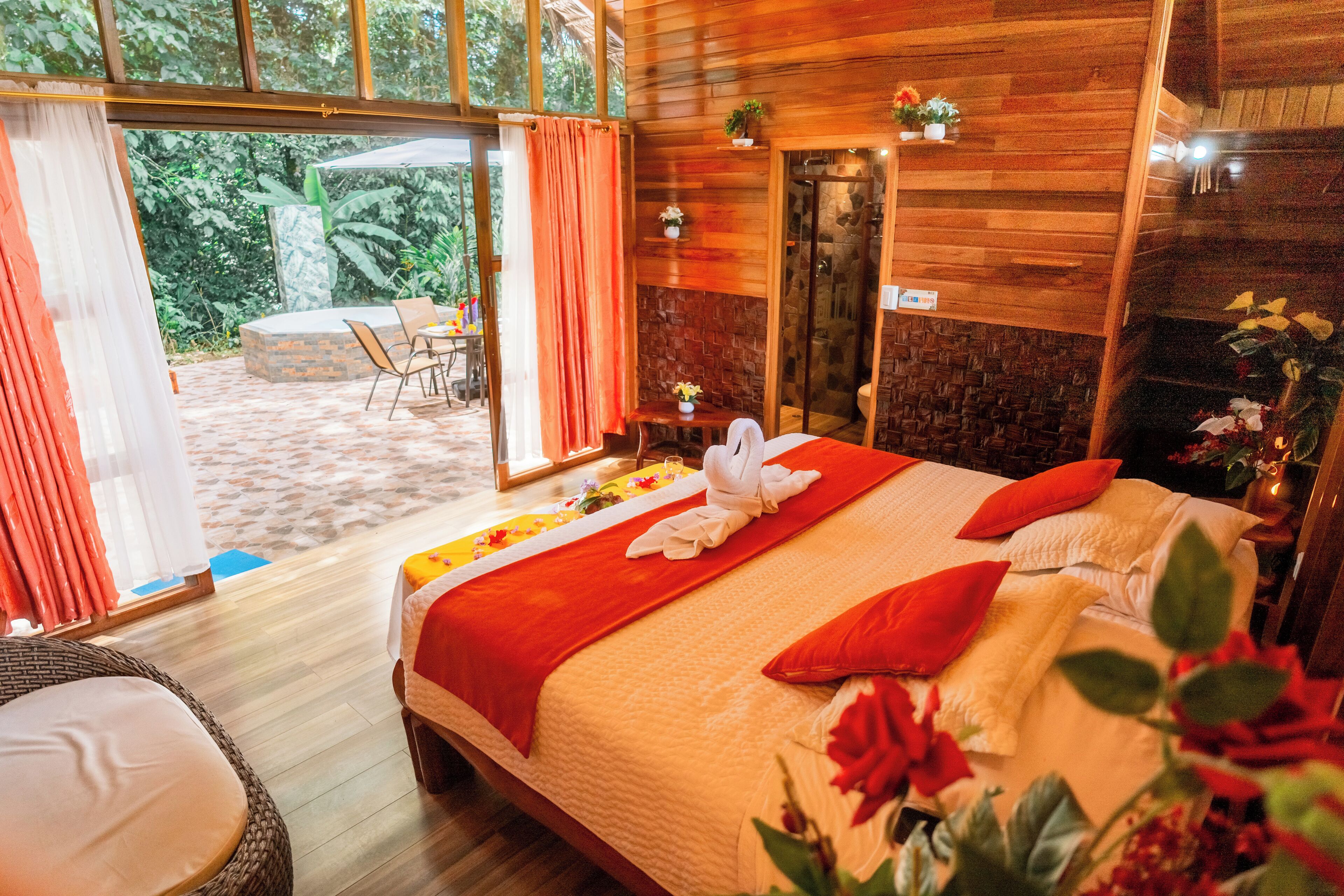 Una habitación amplia con una cama grande, piso de madera, puerta corredera de vidrio que da a un área exterior, y una mesita con una silla.