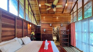 Una habitación de madera con una cama grande, un ventilador de techo y un baño visible a través de una puerta de cristal.