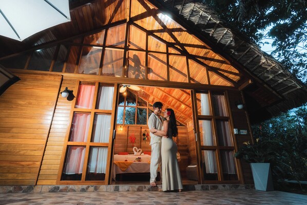 A wooden cabin with a thatched roof, large windows, and a bed with a white sheet and red curtains.