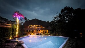 Un área de piscina con un edificio de techo de paja, un sendero de piedra e iluminación decorativa.