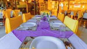 Un restaurante con una larga mesa dispuesta para comer, sillas amarillas y un mantel morado.