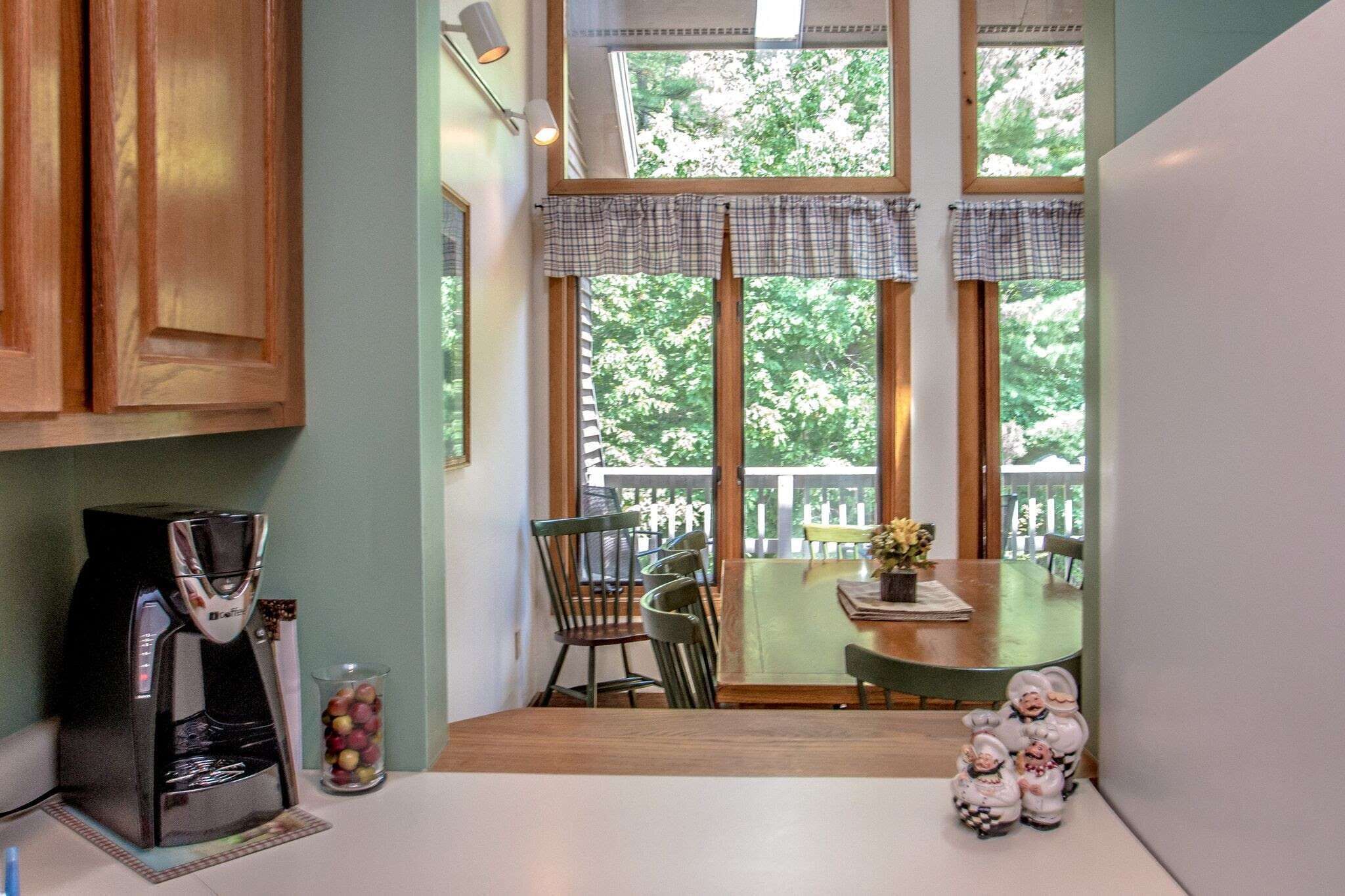 A kitchen with a coffee maker, a glass of fruit, and a dining area with chairs and a table.