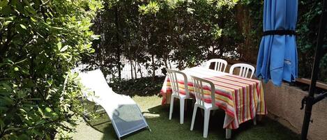 A patio area with a table and chairs, surrounded by greenery and a blue umbrella.