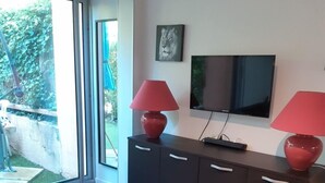 A modern living room with a dark wooden cabinet, two red lamps, a flat-screen TV, and a sliding glass door leading to a garden. - Studio with private garden (Sanary-sur-Mer)