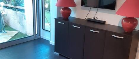 A modern living room with a dark wooden cabinet, two red lamps, a flat-screen TV, and a sliding glass door leading to a garden.