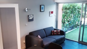A modern living room with a dark sofa, a dining table, and a large window offering a view of a garden. - Studio with private garden (Sanary-sur-Mer)