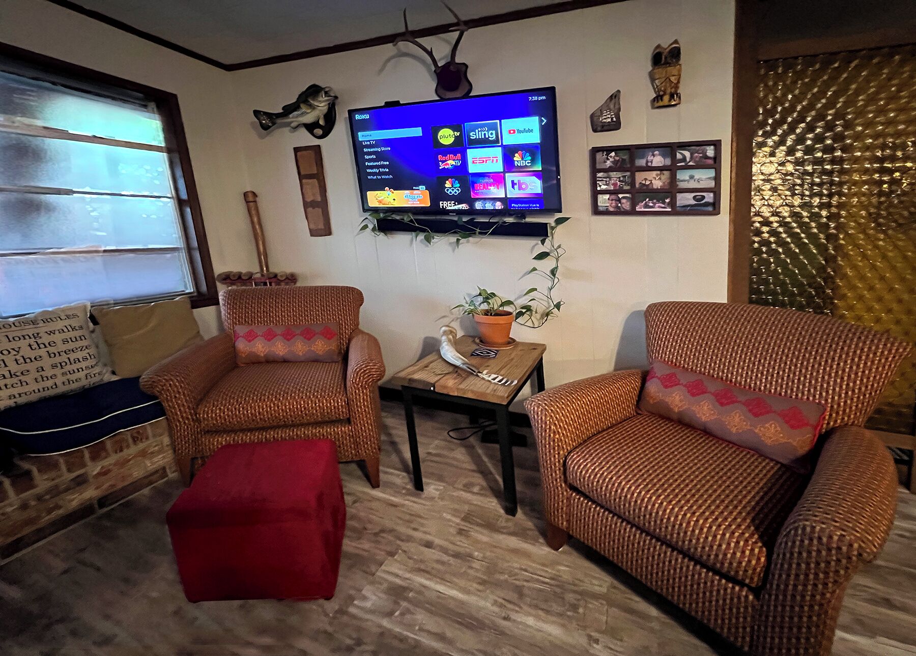 A living room with two wicker armchairs, a red ottoman, a small table with a plant, and a flat-screen TV displaying a streaming service interface.