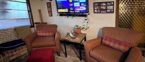A living room with two wicker armchairs, a red ottoman, a small table with a plant, and a flat-screen TV displaying a streaming service interface.
