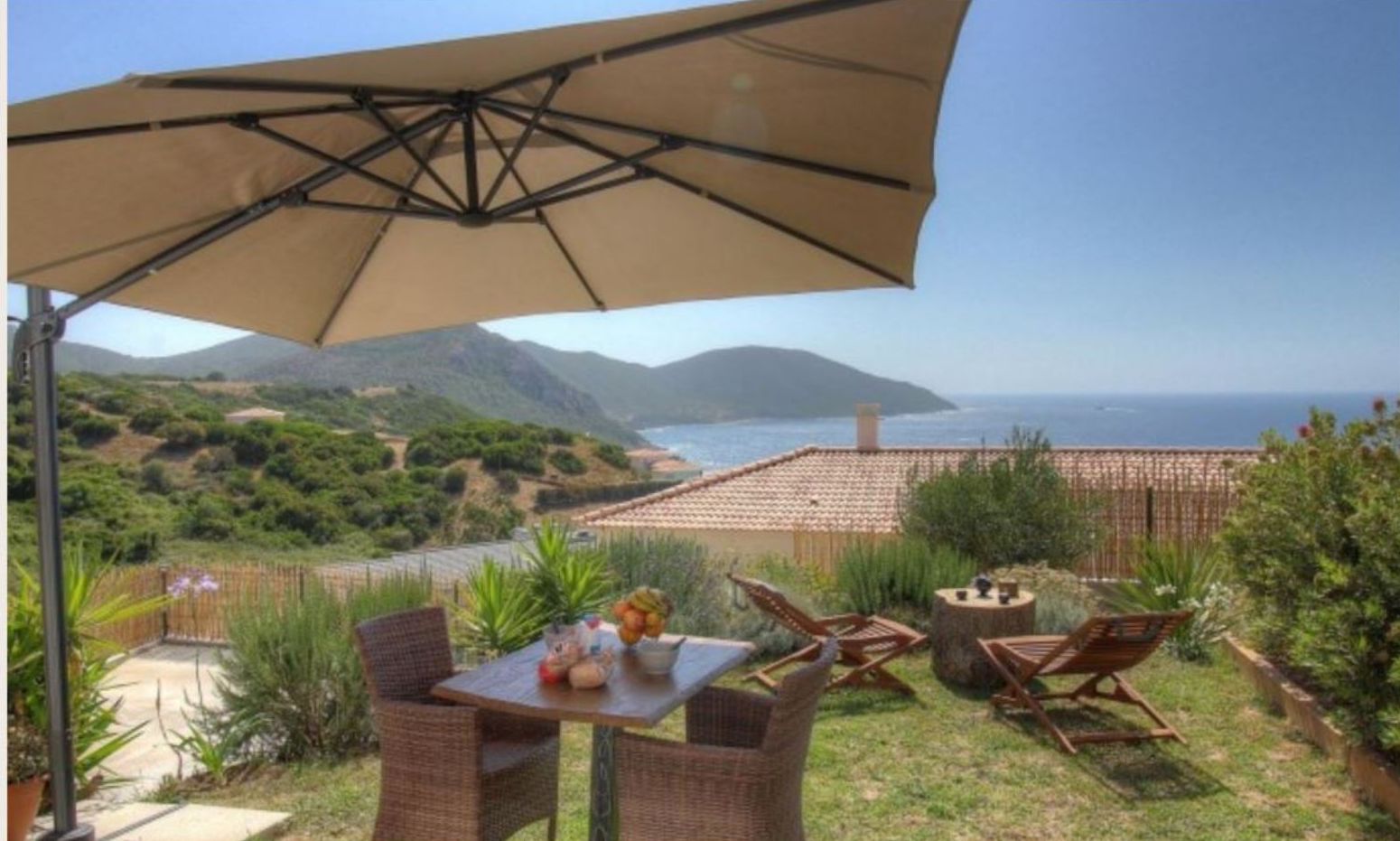 An outdoor patio with a table, chairs, and a large umbrella overlooking a scenic view of the sea and hills.