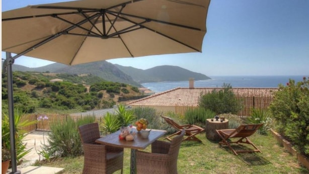 An outdoor patio with a table, chairs, and a large umbrella overlooking a scenic view of the sea and hills.