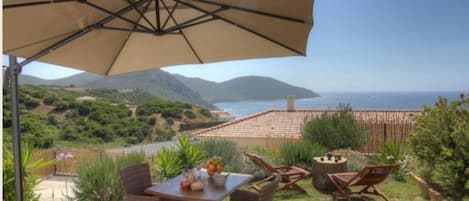 An outdoor patio with a table, chairs, and a large umbrella overlooking a scenic view of the sea and hills.