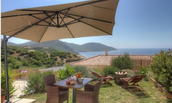 An outdoor patio with a table, chairs, and a large umbrella overlooking a scenic view of the sea and hills.