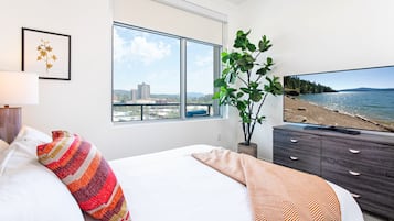 A bedroom with a large window, a bed with striped pillows, a television displaying a beach scene, and a potted plant.