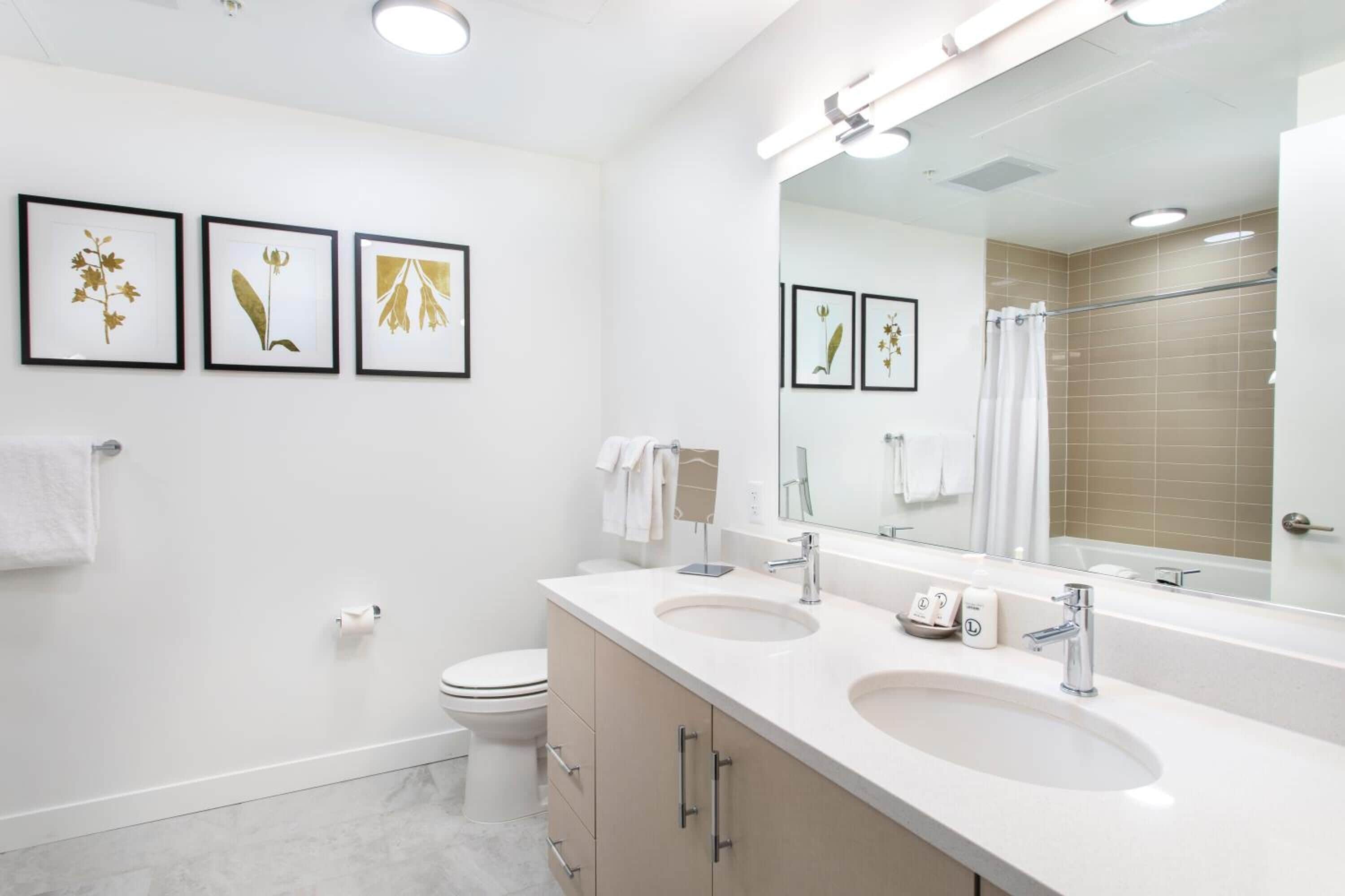 A modern bathroom with two sinks, a toilet, a large mirror, and framed botanical artwork on the wall.